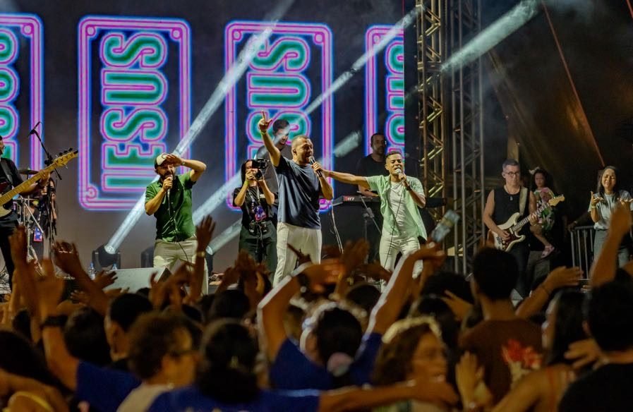 Público de mãos erguidas diante do palco com painéis luminosos escritos Jesus