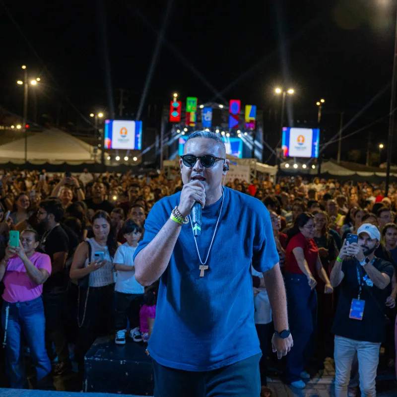 Cantor de óculos escuros ao microfone no palco, com a multidão lotando o Parque Shopping Maceió atrás.