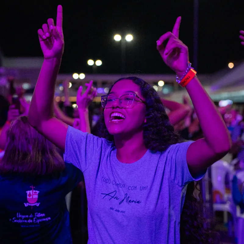 Jovem de braços erguidos e sorriso durante o louvor, sob a luz roxa do Luau.