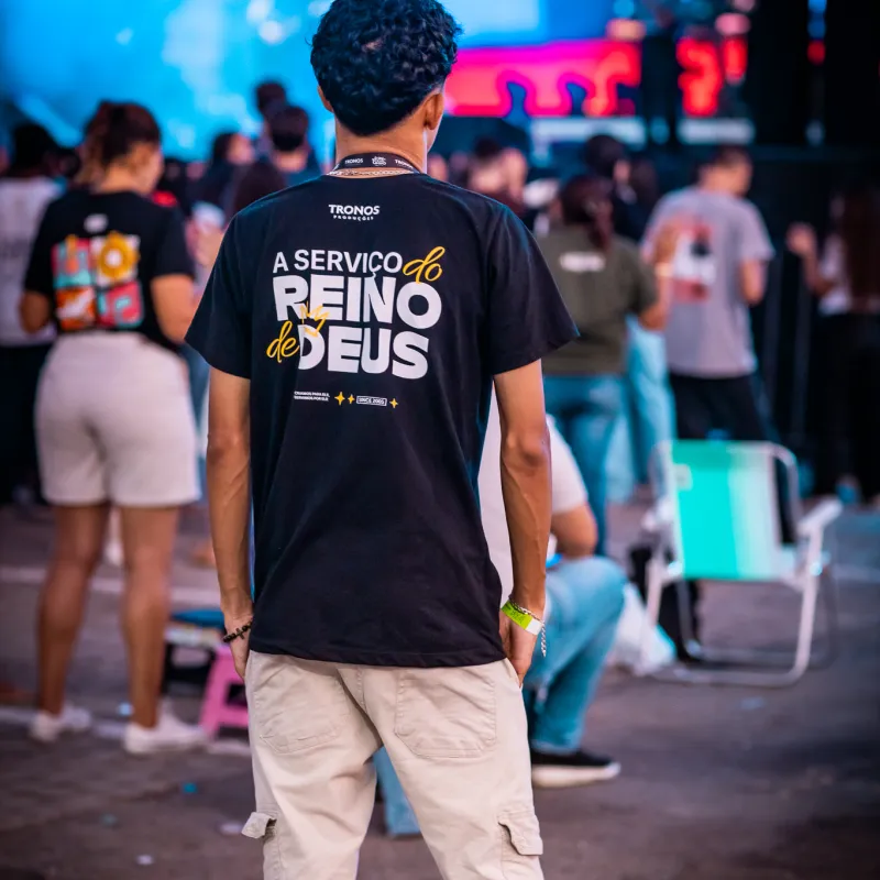 Jovem de costas na plateia com a camiseta da Tronos Produções escrita "A serviço do Reino de Deus".