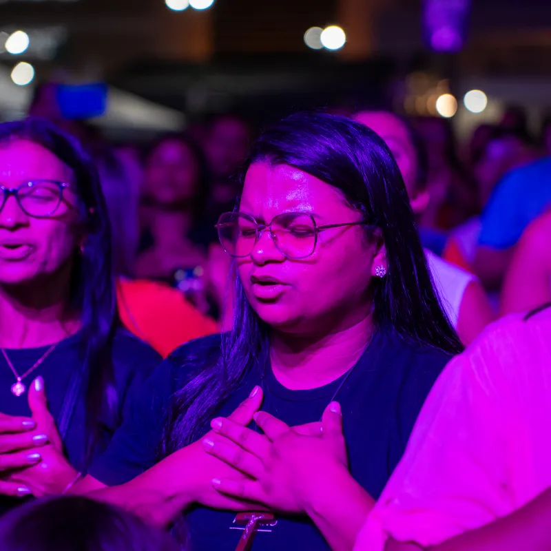 Mulher de olhos fechados e mãos sobre o peito, em oração, sob a luz roxa do Luau.