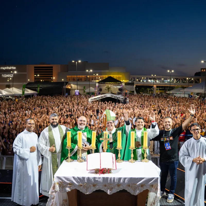 Padres e o bispo diante do altar no palco, com a multidão de braços erguidos ao fundo, no Parque Shopping Maceió.