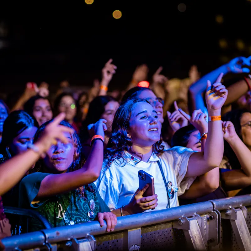 Público junto à grade com as mãos erguidas sob a luz azul, diante do palco do Luau.