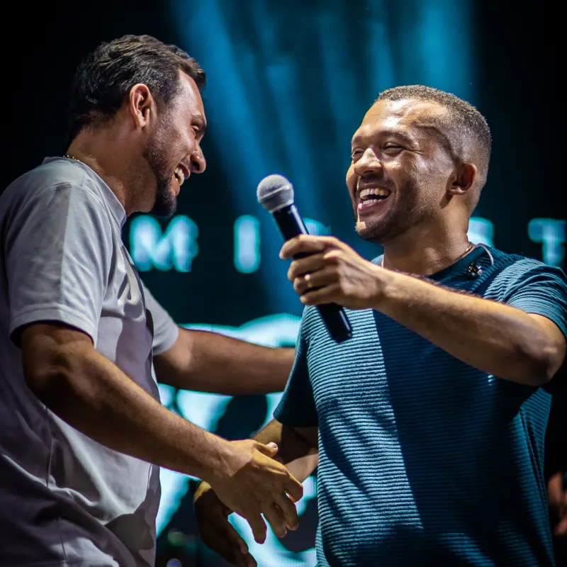 Dois homens sorrindo e se cumprimentando no palco do Luau, um deles ao microfone.